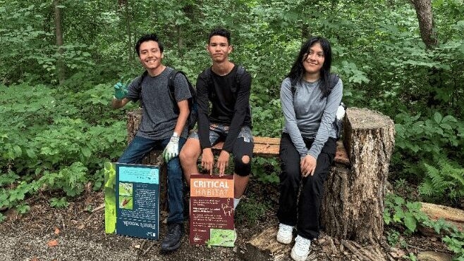 Three youths sit on a bench in the woods holding signage