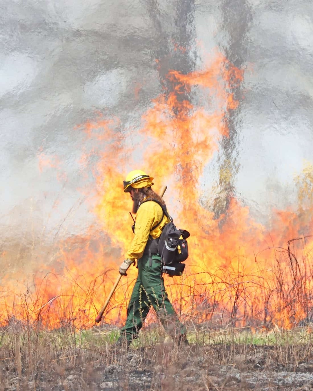 a man in protective fire gear with a handheld tool and a backpack in front of a large flame