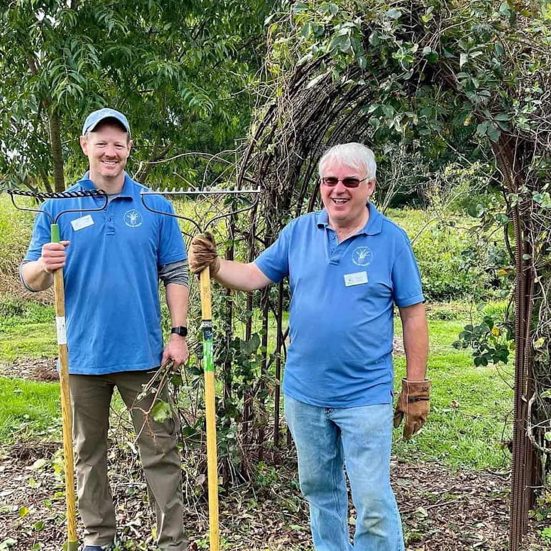 Two men in bright blue shirts smile at the camera holding garden tools.