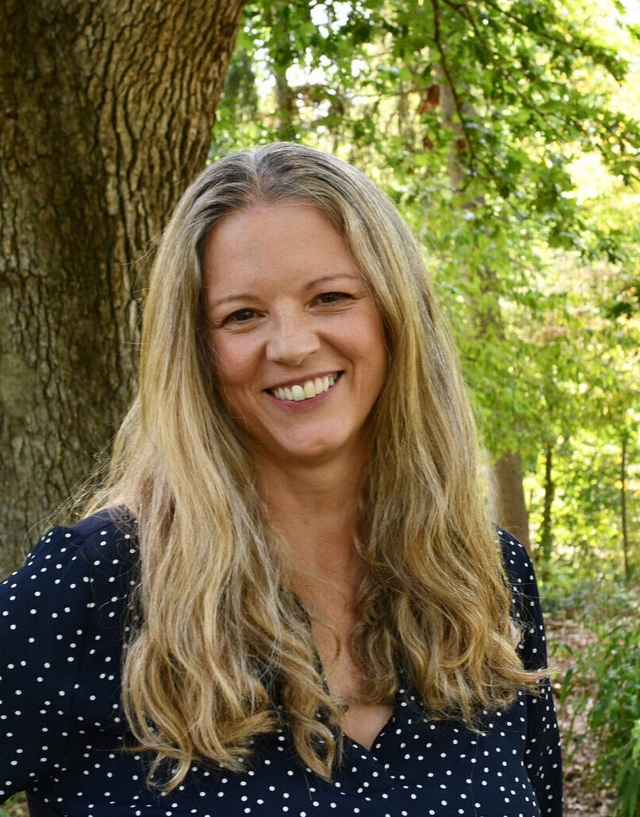 A woman with blonde hair and a blue polka dot shirt smiles at the camera.