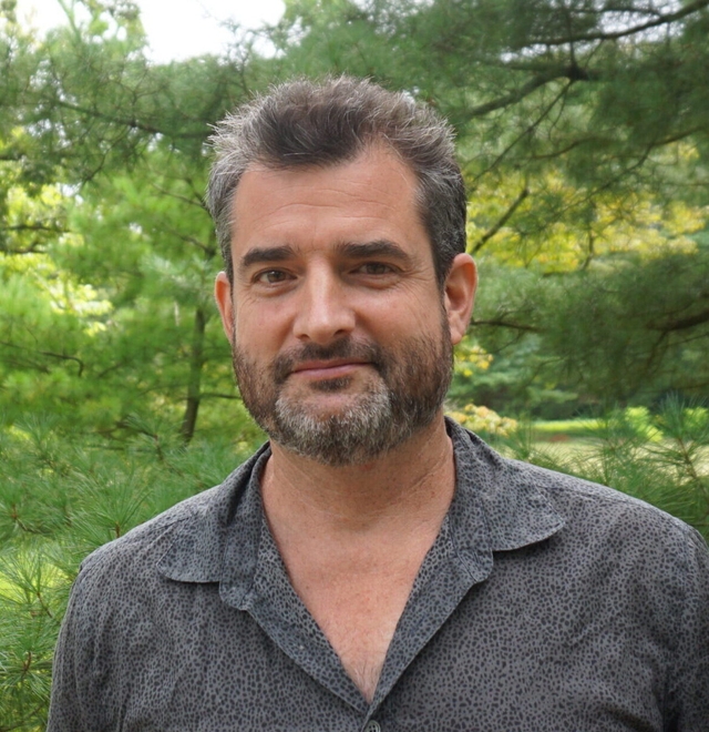 A man in a dark gray shirt with a nametag on it smiles at the camera.