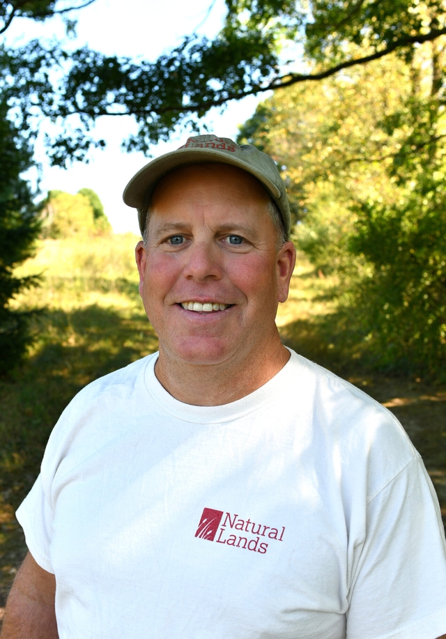 A man in a baseball cap and white Natural Lands tshirt smiles at the camera