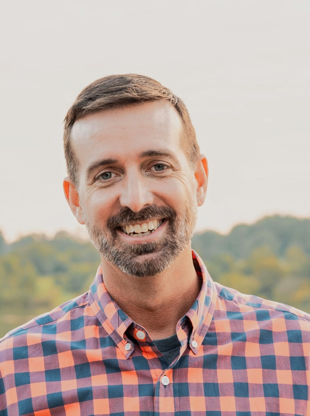 A man with a short beard and a plaid shirt smiles at the camera.