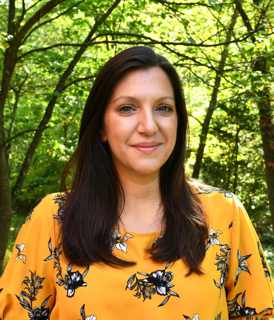 A woman with long brown hair and a yellow shirt looks at the camera