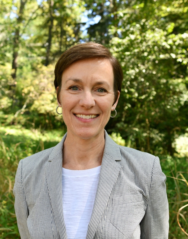 A woman with short brown hair and a gray jacket smiles at the camera.