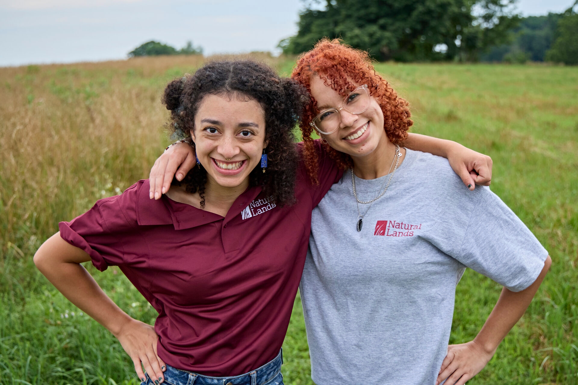 Two young women of color stand in Natural Lands logo shirts with their arms around each other and smile at the camera.