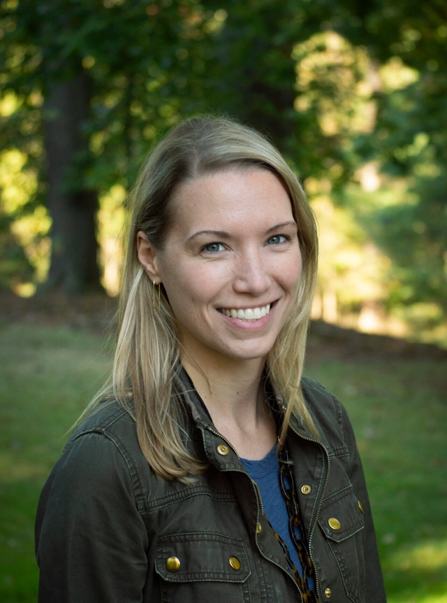 A smiling woman with blond hair wearing a black jeans jacket looking at the camera with trees and a hillside in the background