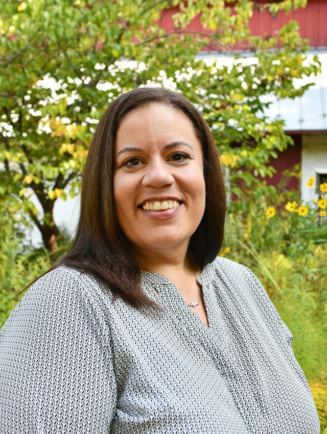 A smiling woman wearing a patterned shirt looking at the camera with a barn and yellow flowers in the background.