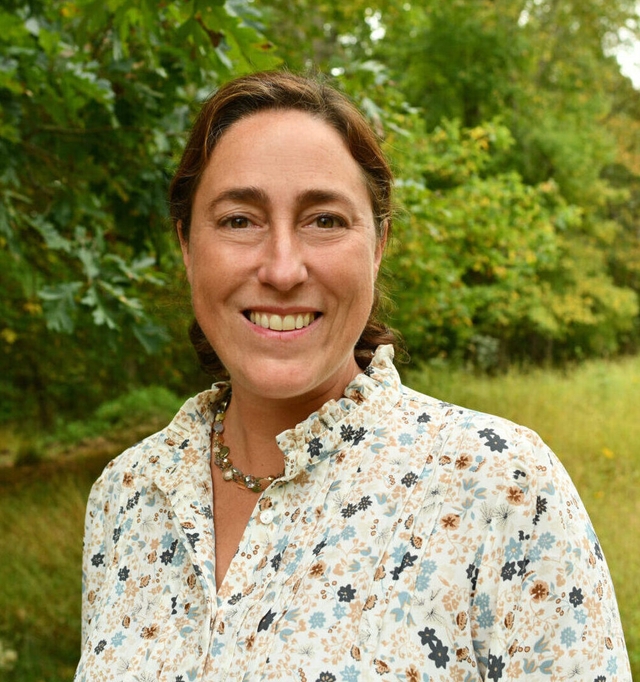 a smiling woman wearing a necklace and floral-print shirt standing in front of a meadow and trees