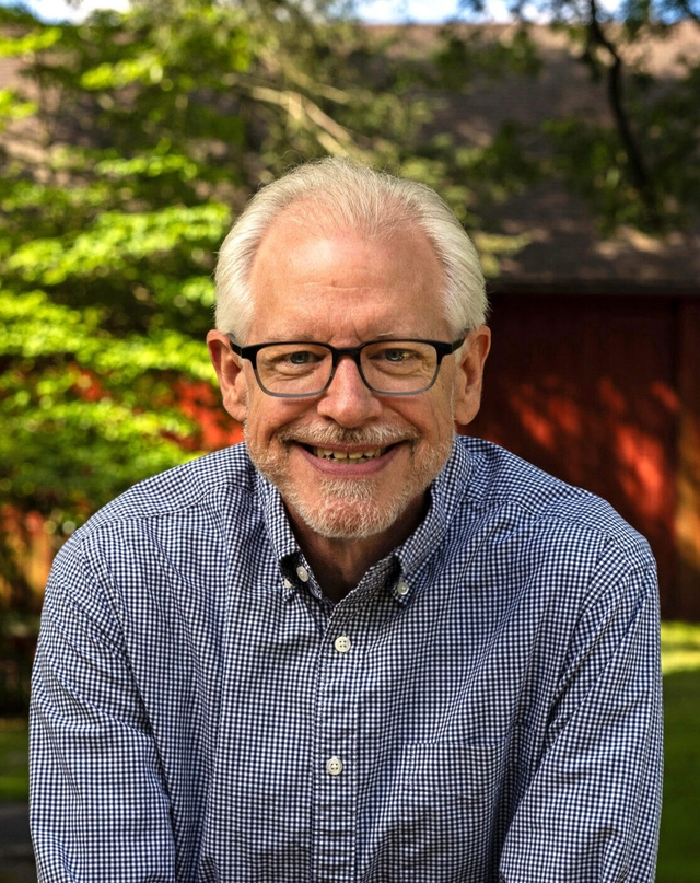 a smiling man with white hair and beard wearing glasses and a blue and white checkered shirt leaning on a stone wall with a barn in the background