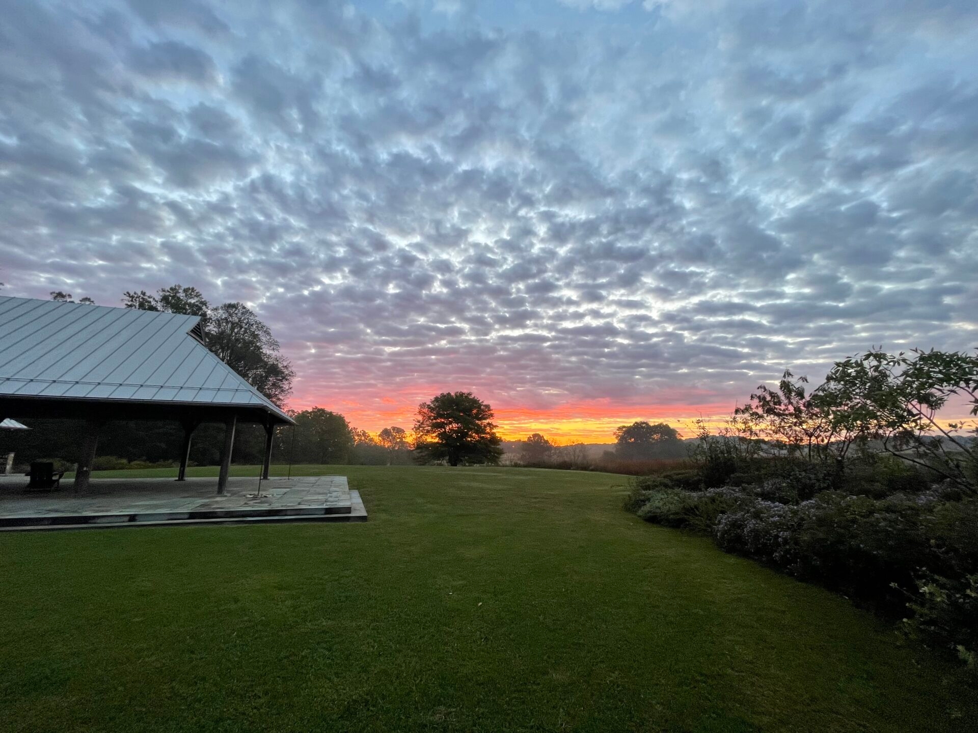 dawn and a cloud-filled sky with a pavilion, lawn, trees, and plantings. - Dawn and rolling clouds at the Lenfest Center at ChesLen Preserve in summer. 
