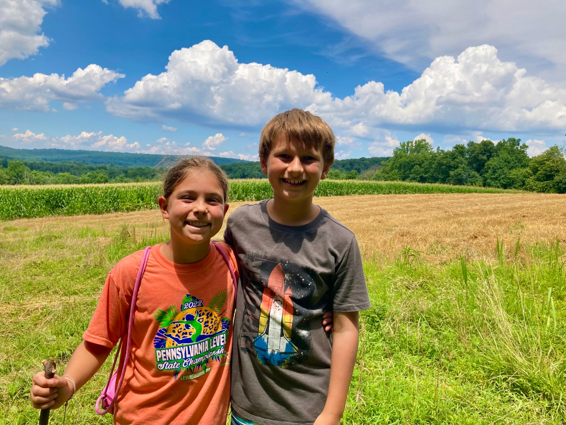 a young boy and girl standing with arms around each other smiling, one with a walking stick, with an agricultural field and blue sky with fluffy white clouds in the background - Two kids from Nature Camp enjoying a hike through the agricultural fields at Crow’s Nest Preserve in summer.
