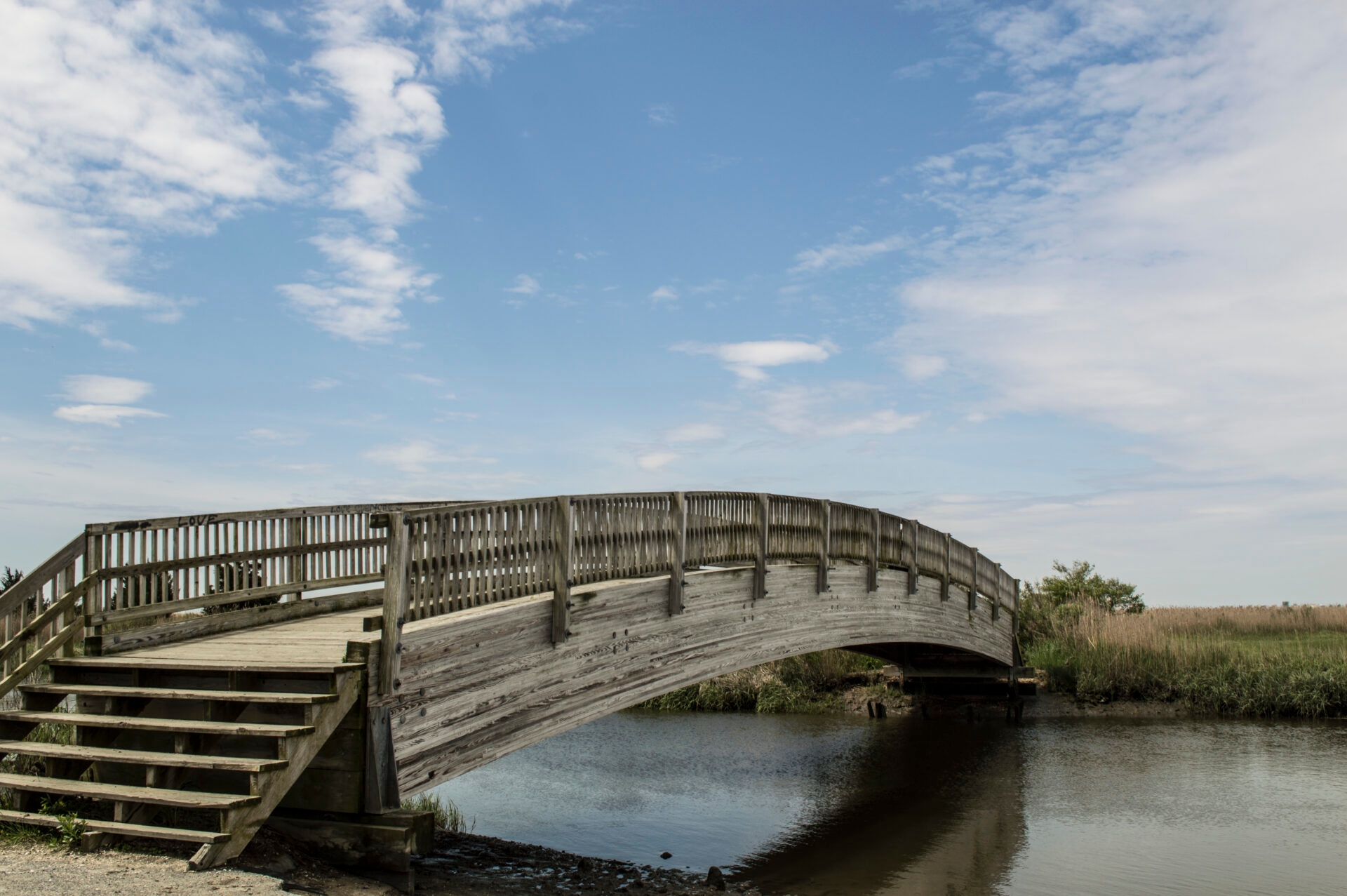 a low, arching, wooden bridge over water with a blue sky and some white clouds - The bridge on the Turkey Point Water Trail with a blue sky at Glades Wildlife Refuge, NJ.
