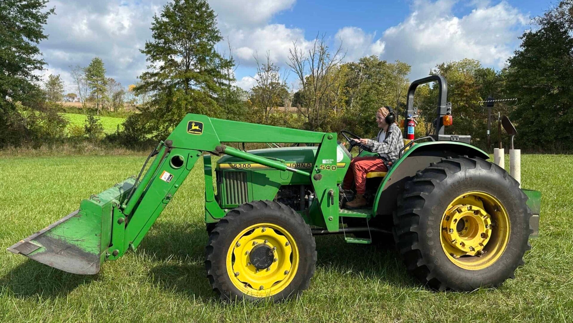 A woman driving a green John Deere tractor outside in a field on a sunny day.