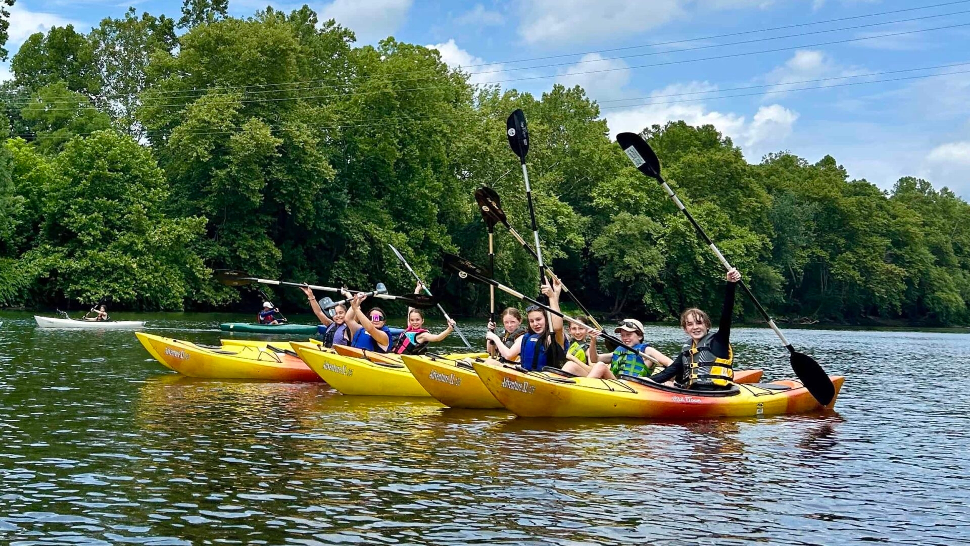 Kids in yellow kayaks on a river holding oars up with joy