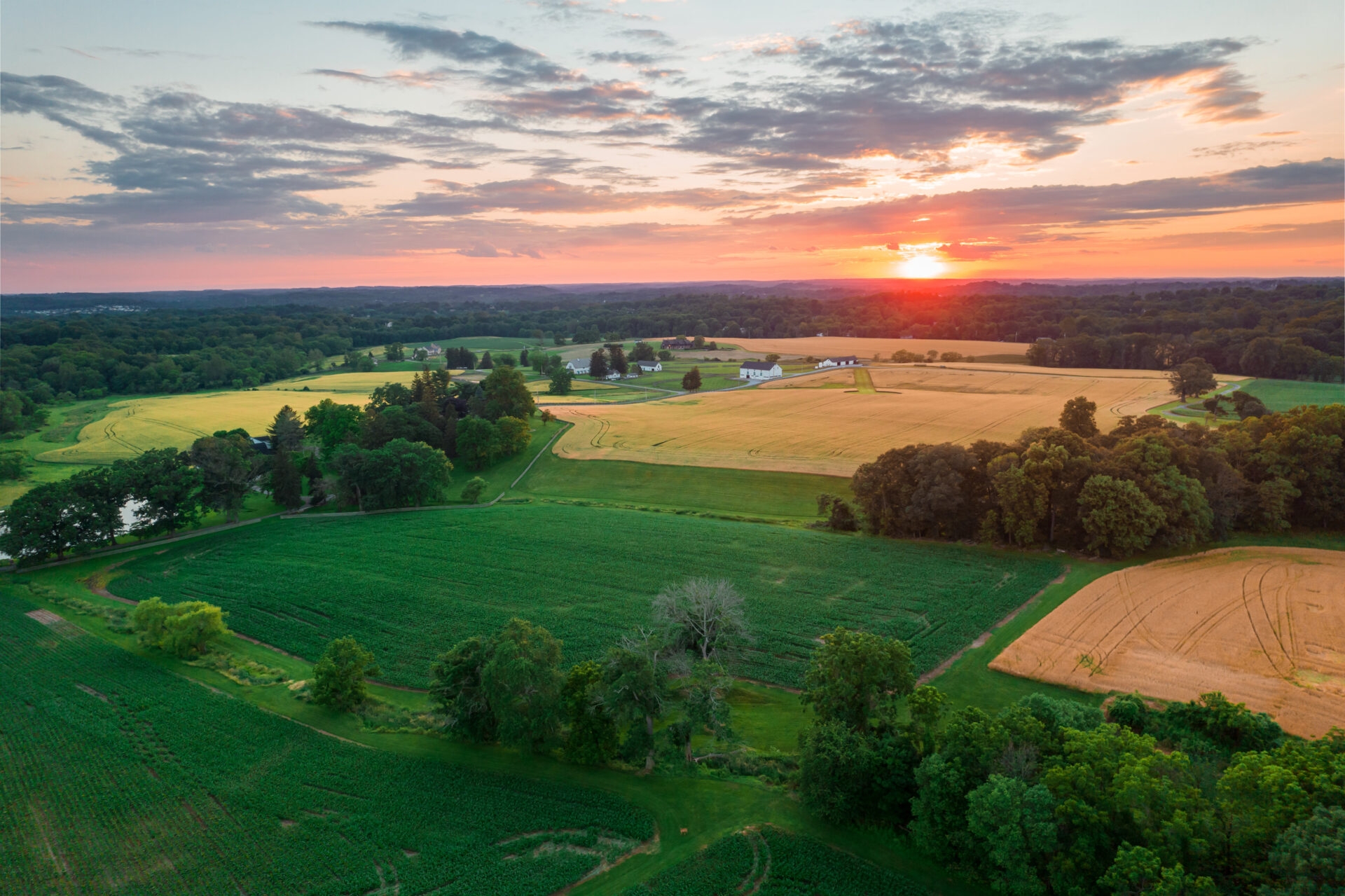 an aerial view of an orangish sunset over a vast landscape of green trees and green and brown agricultural fields.