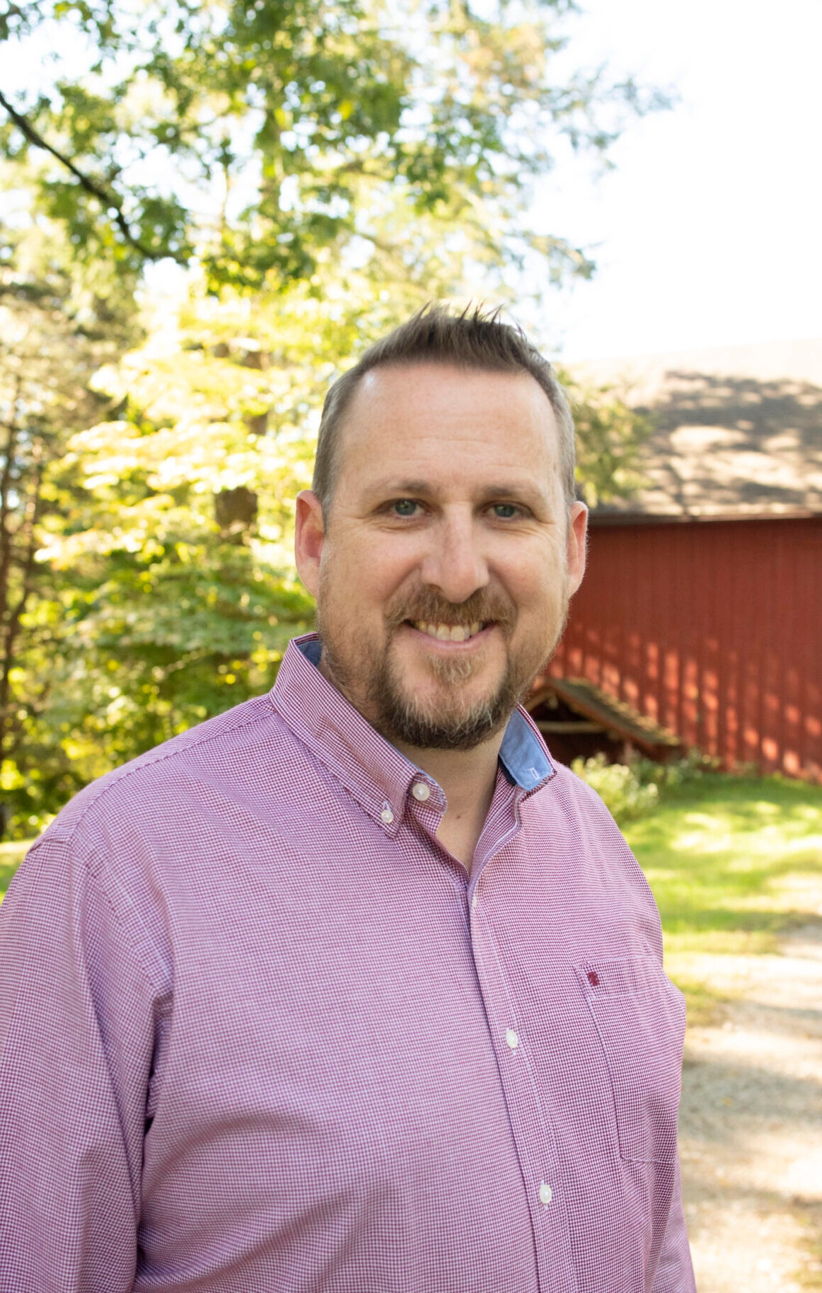 a smiling man with beard and mustache wearing a red and white checkered shirt with a red barn in the background