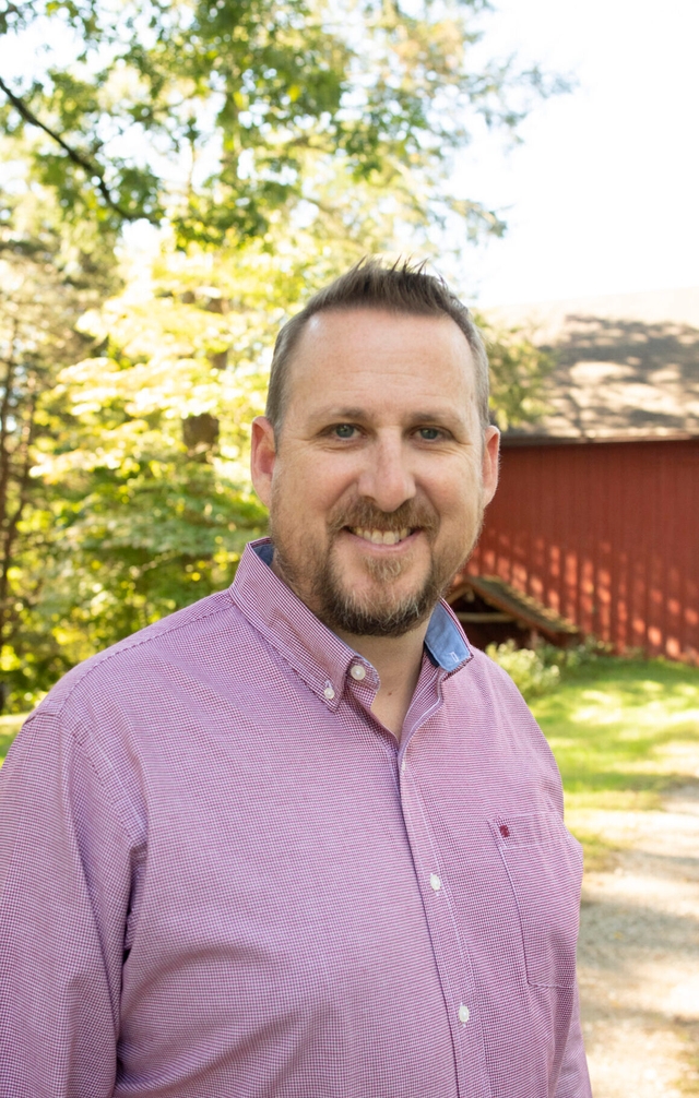 a smiling man with beard and mustache wearing a red and white checkered shirt with a red barn in the background