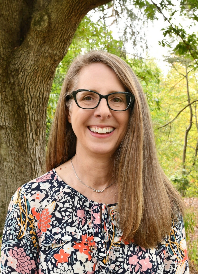 a smiling woman with glasses, a necklace, and a floral patterned shirt in front of a large tree and forest