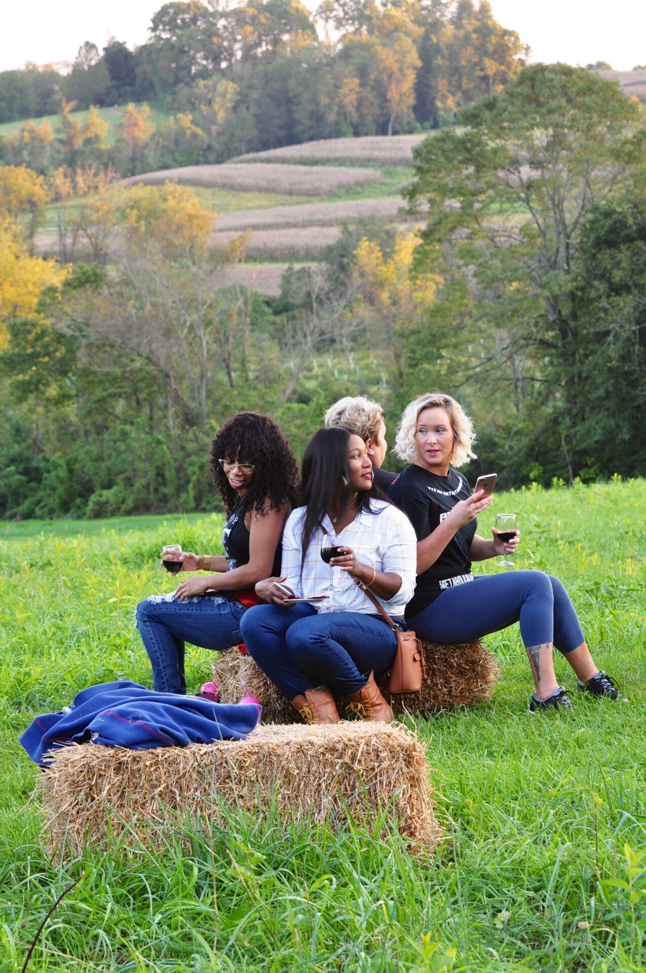 four women sitting on a hay bale in a green field sipping wine from glasses