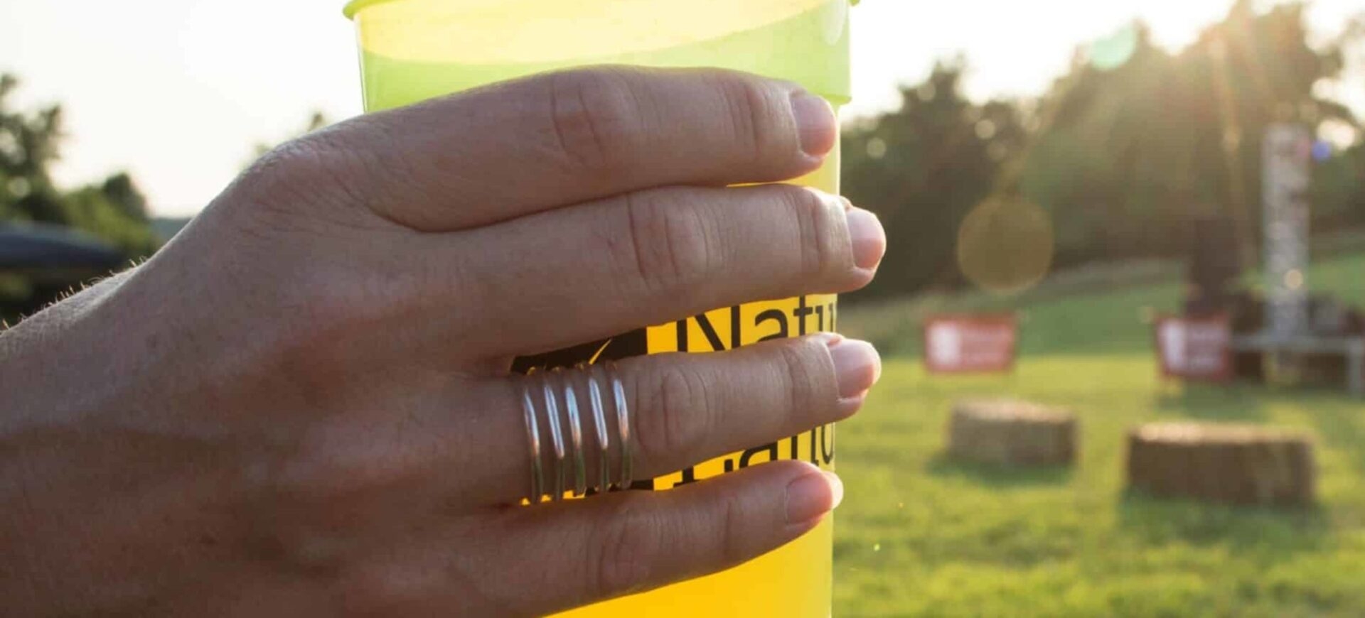 a hand with multiple rings on it holding a natural lands cup with a grassy background