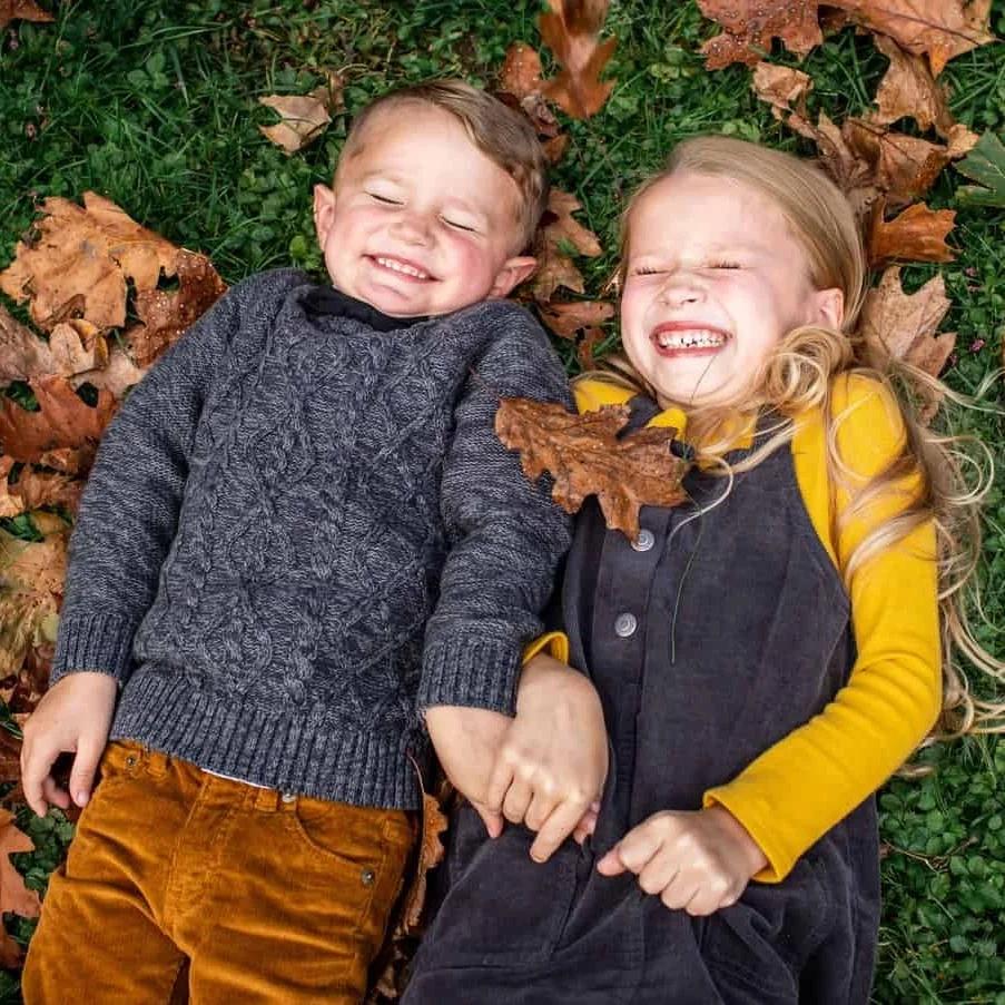 A girl and a boy in fall clothing smile with their eyes closed while lying on green grass and brown autumn leaves.