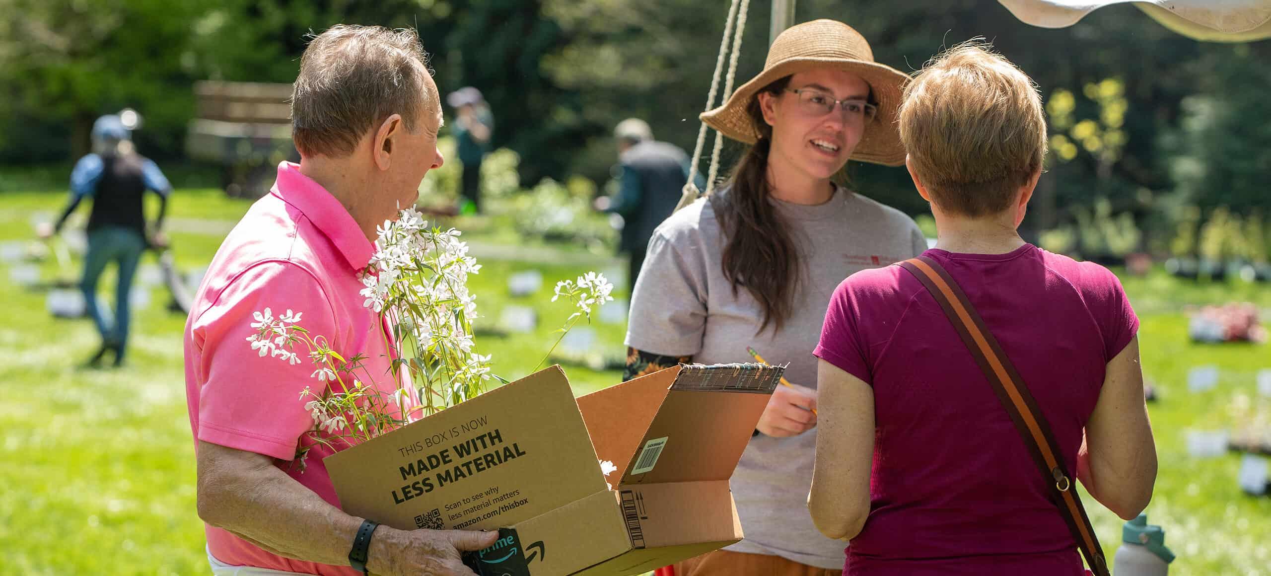 a man carries a box of blooming plants and two women in summer clothes speak to each other