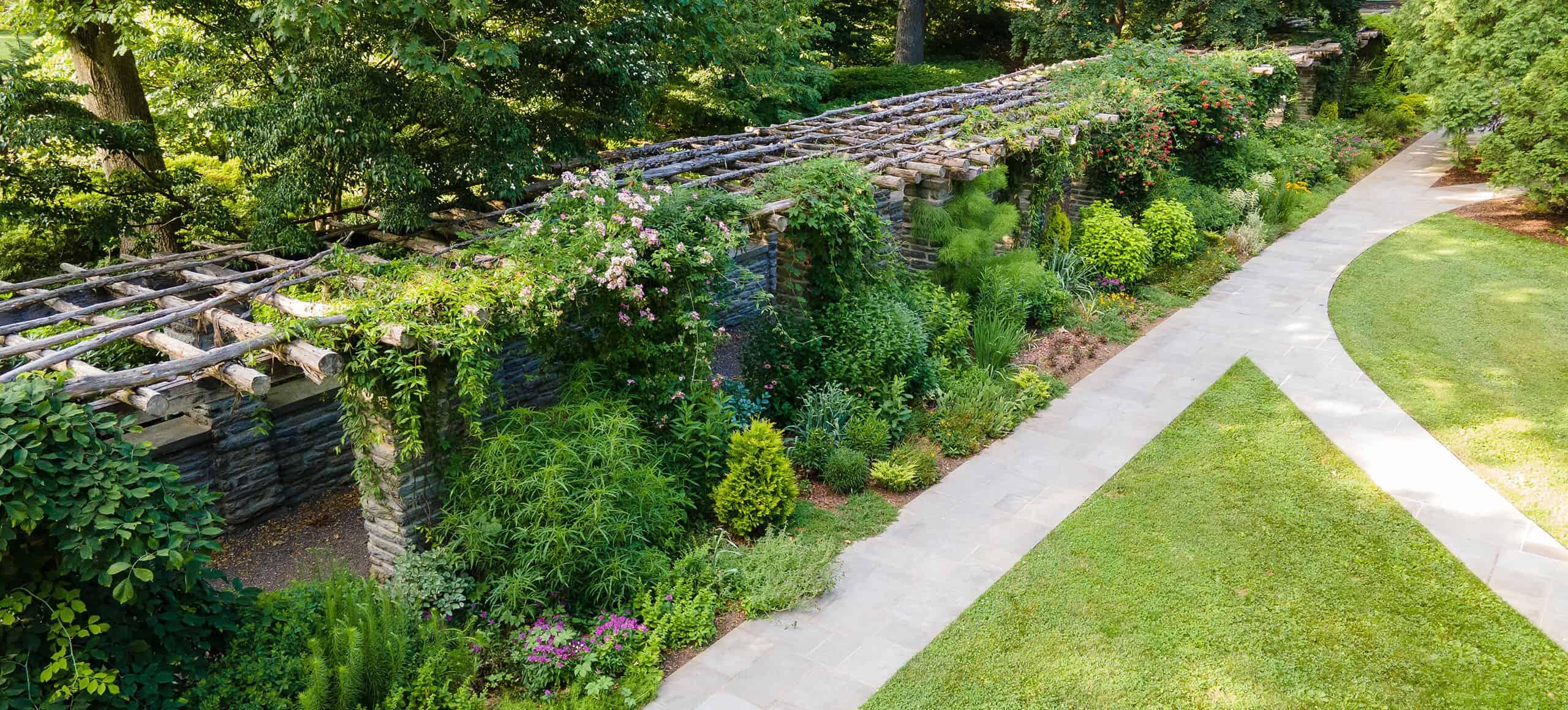 drone photo of large stone pergola with wooden poles on top, lushly planted