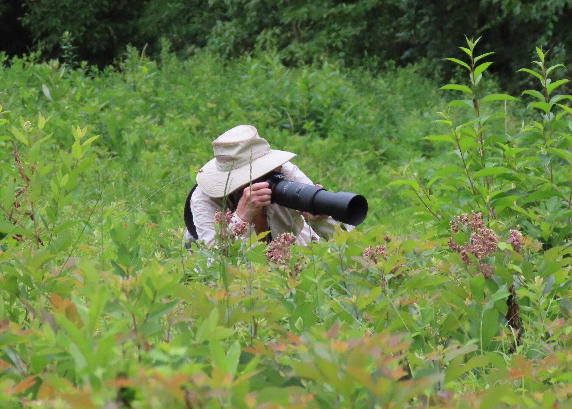 a photographer with a hat and a long camera lens in a green meadow taking photos