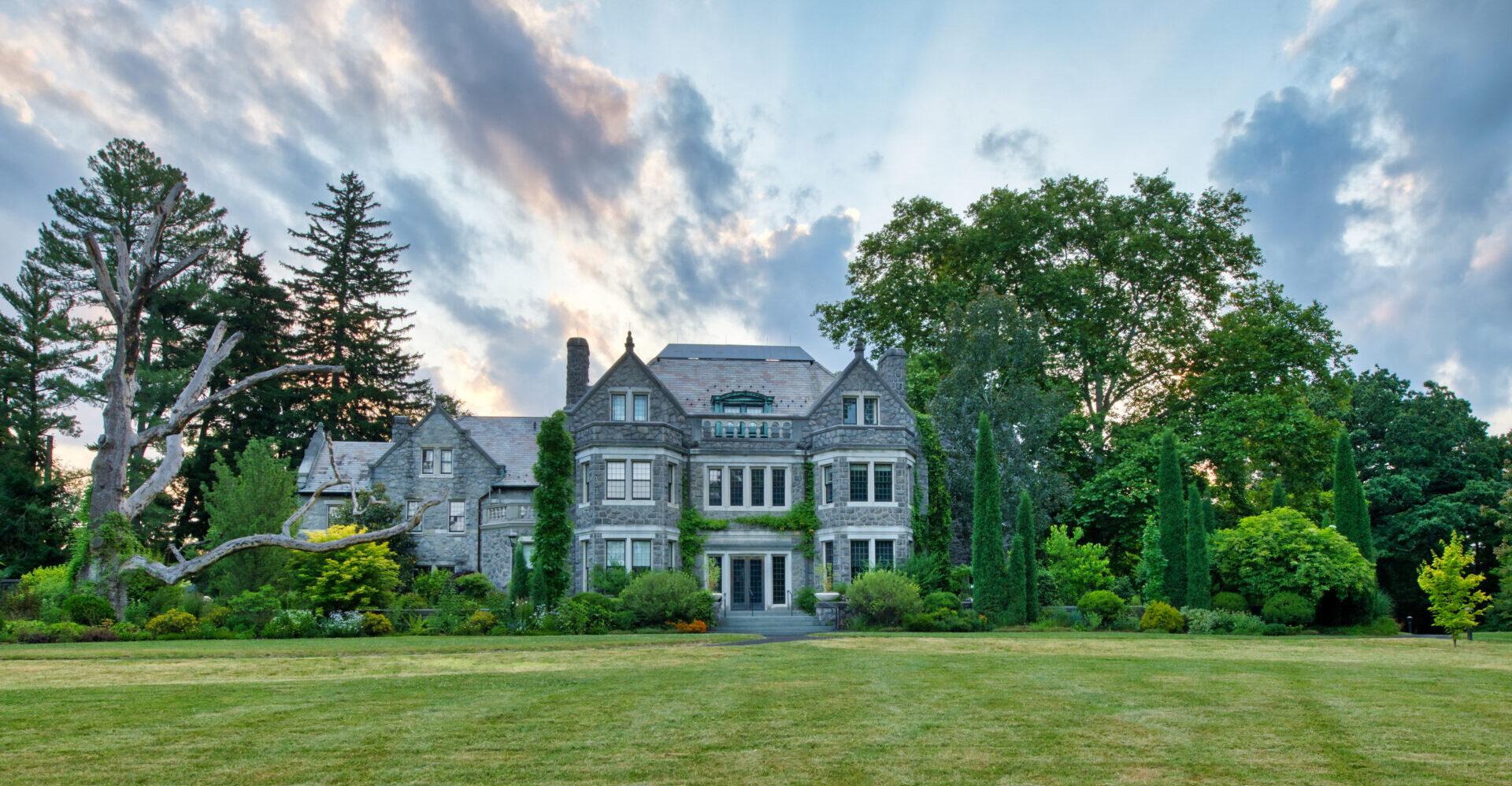 a large stone mansion surrounded by trees and shrubs with a vast green lawn and blue and pinkish clouds in the sky