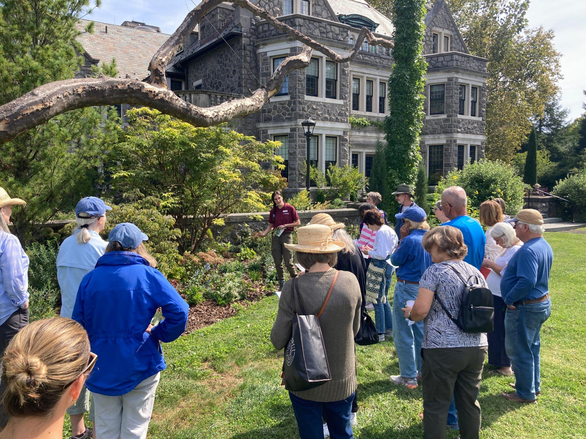 a group of people crowded around a woman who is pointing to a garden bed of plantings with a large stone house in the background