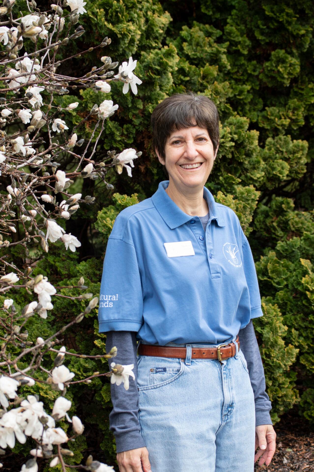 a woman smiling wearing a blue shirt and jeans standing near a shrub with white flowers