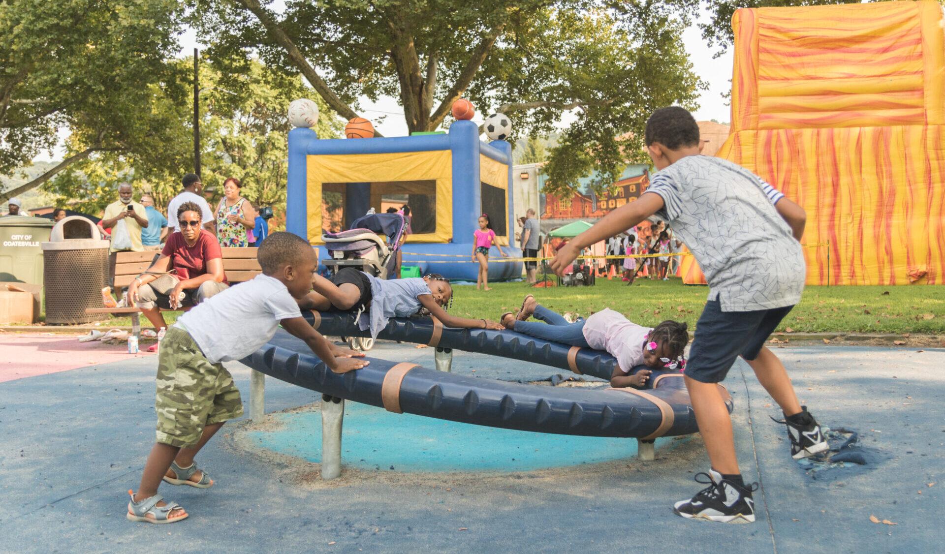 four children playing on a round playground ride at an outdoor celebration in a park