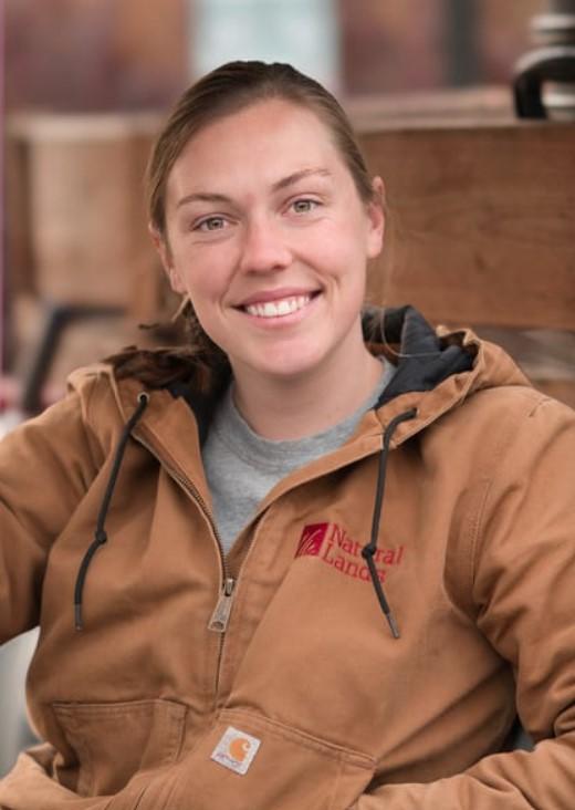 A young woman in a brown Natural Lands coat smiles at the camera.