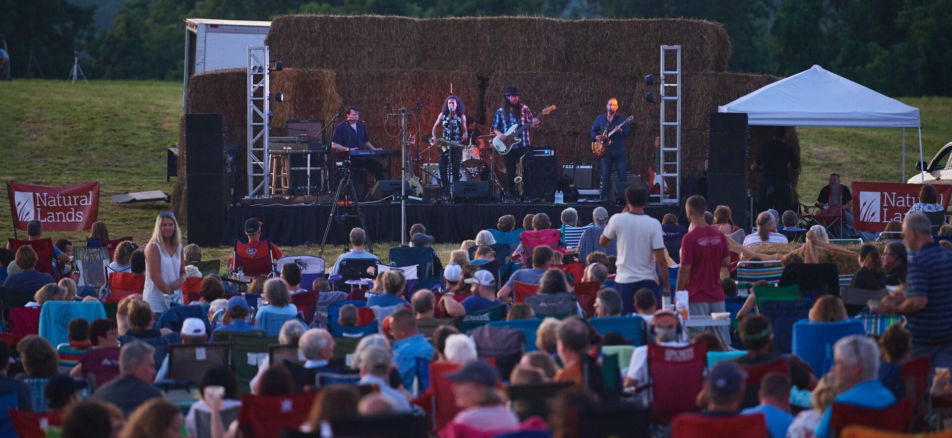 A band plays in front of a stack of hay bales in front of a crowd sitting on camp chairs in twilight outdoors