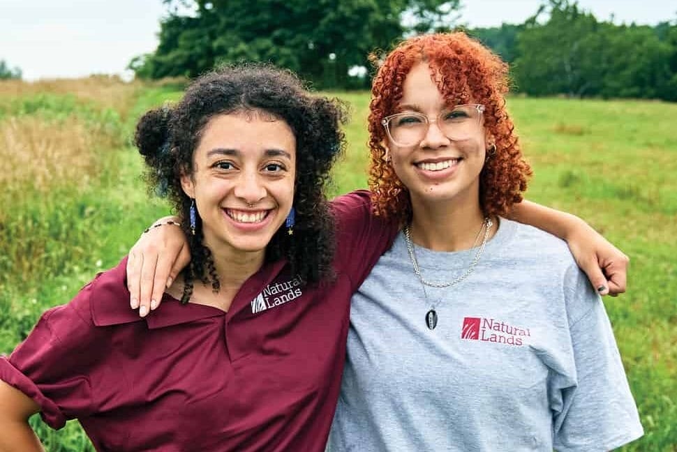 Two young women stand with arms around each others' shoulders in a green meadow