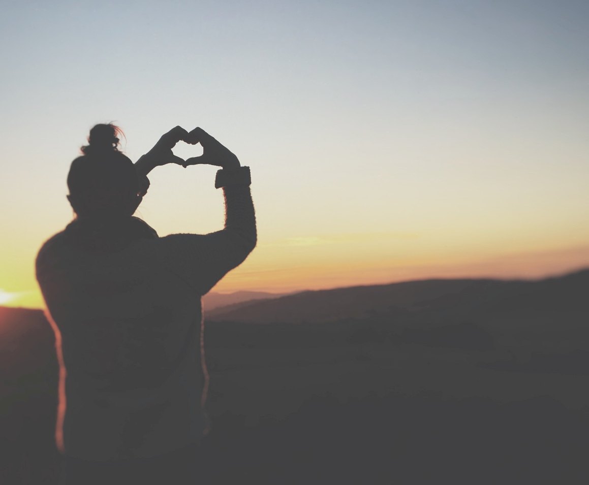 A silhouetted person faces away, looking toward an orange sunrise, and forming the shape of a heart with their hands.
