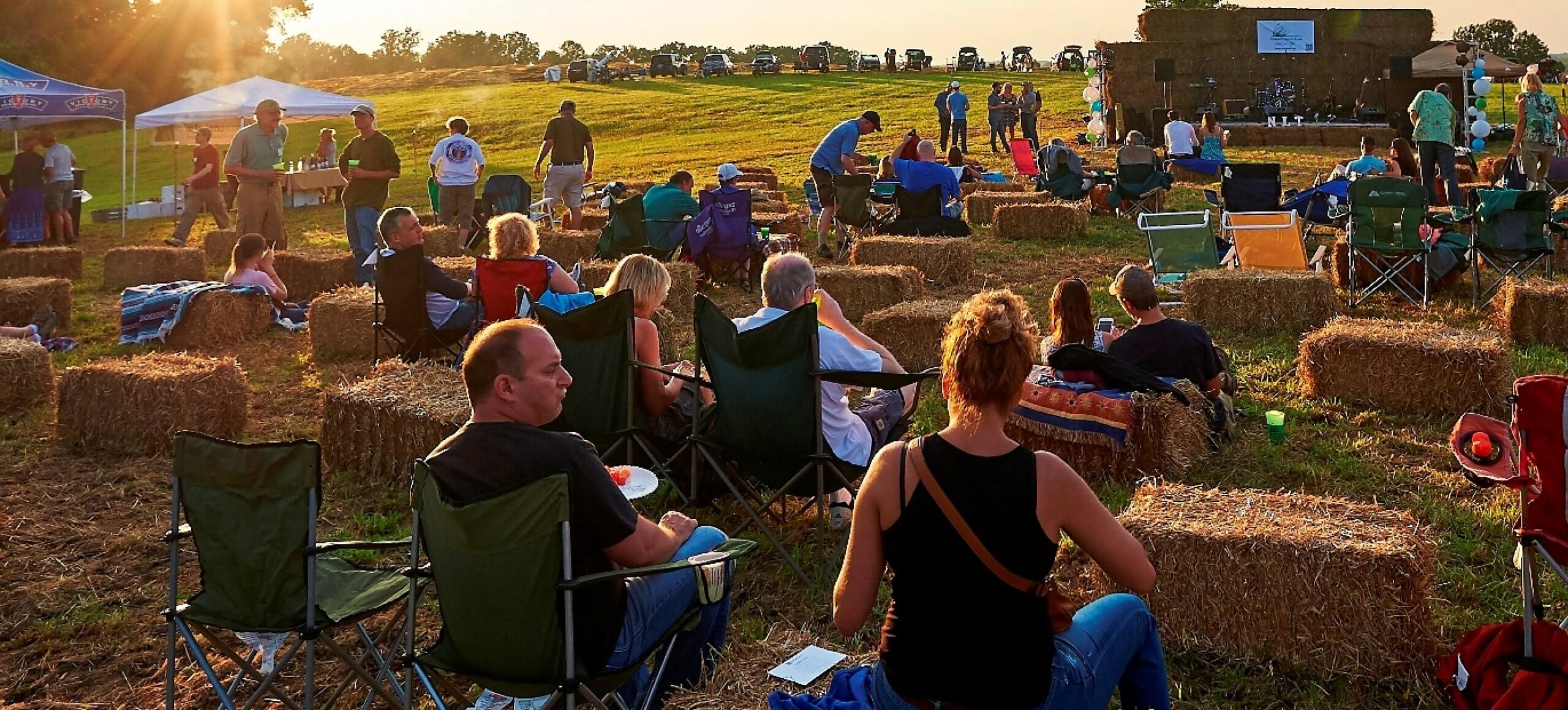 A crowd of people sitting on haybales and camping chairs while the sun goes down.