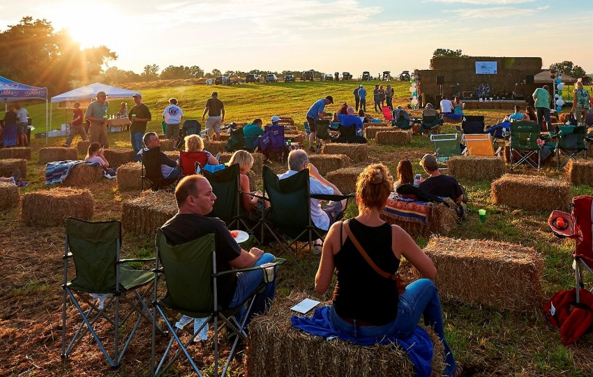 A crowd of people sitting on haybales and camping chairs while the sun goes down.