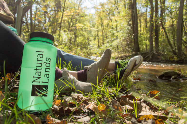 A green water bottle with the Natural Lands logo sits outdoors in front of the legs of two people wearing casual clothes. A creek runs to the right and a forest is in the background.