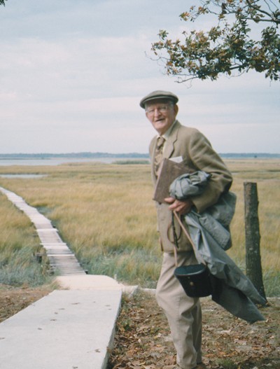 A man holding a coat and bag stands beside a boardwalk trail that leads towards a marsh.
