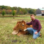 A photo of Dan Barringer outdoors kneeling next to a brown cow.