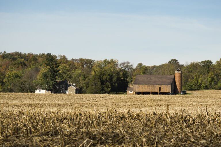A stone house and a barn on a crop field with trees in the background.