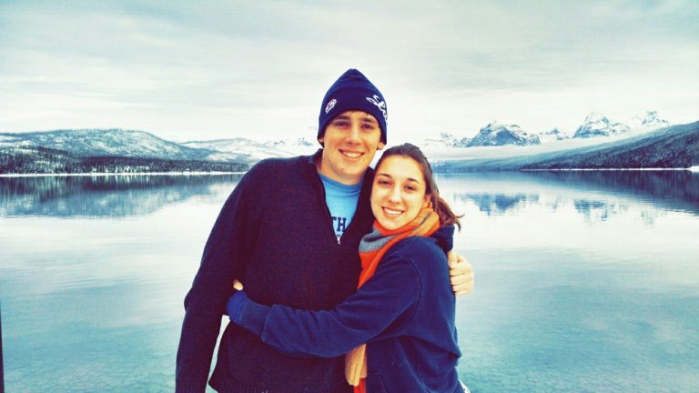 A man and woman hug with a lake and snow covered mountains behind them.