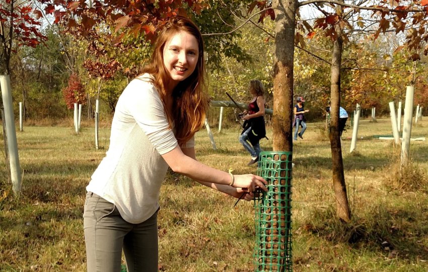 A woman standing next to a tree smiles at the camera in a field of trees with tubes around them.