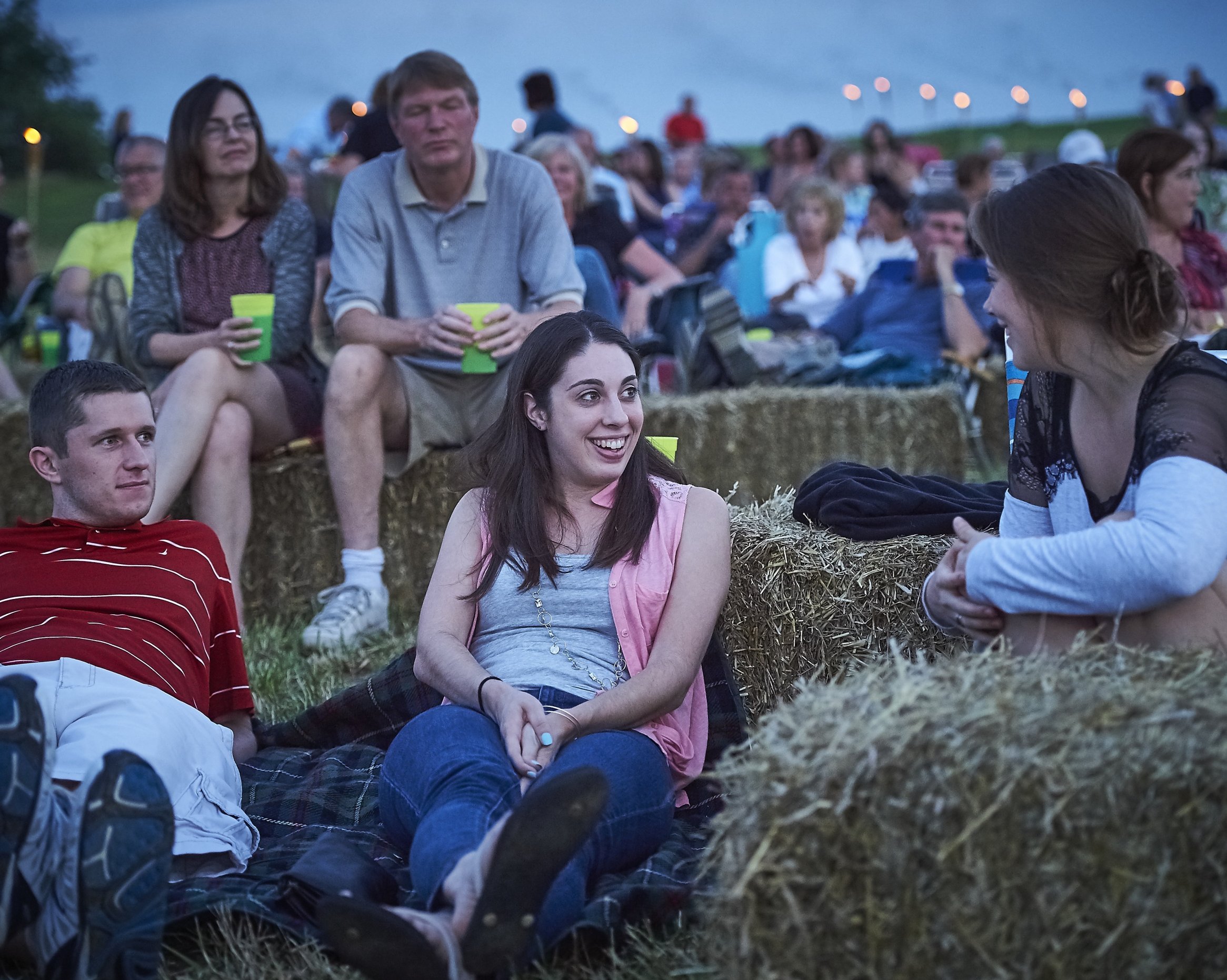 A crowd of people sitting on haybales outdoors in the evening smile and talk.
