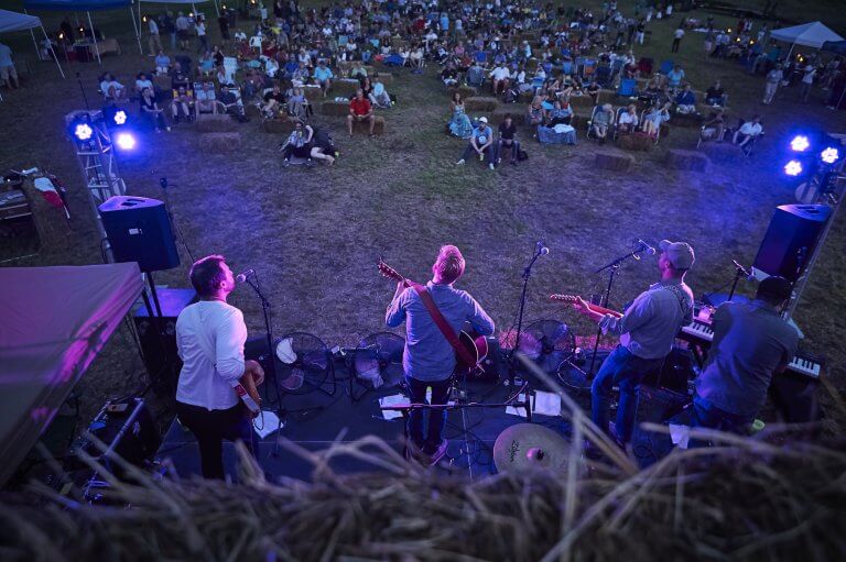 Looking down on a band playing an outdoor concert in front of people sitting on haybales and camping chairs.