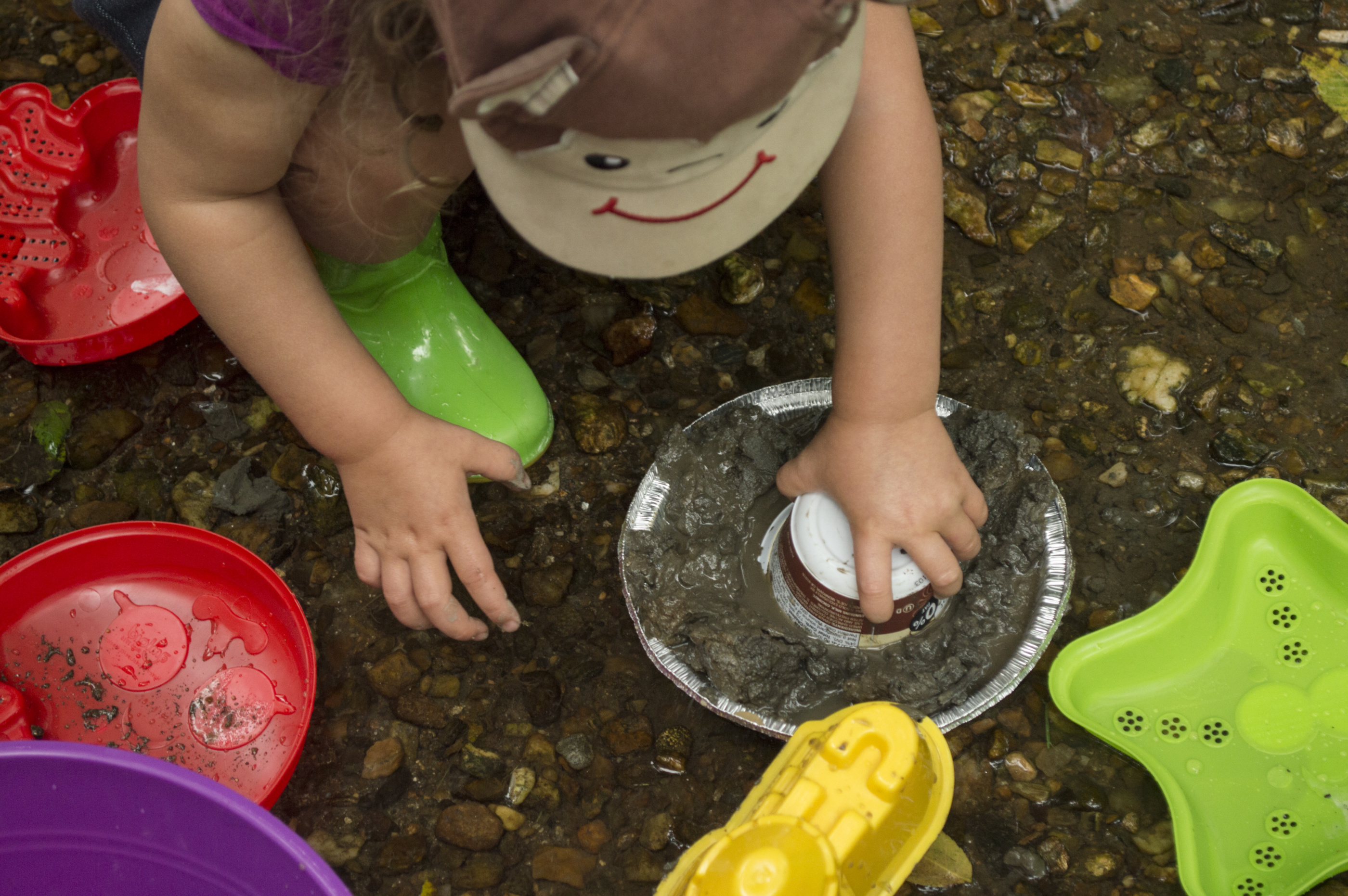 The camera looks down at a child playing in the mud with a plastic cup an tin pie pan.