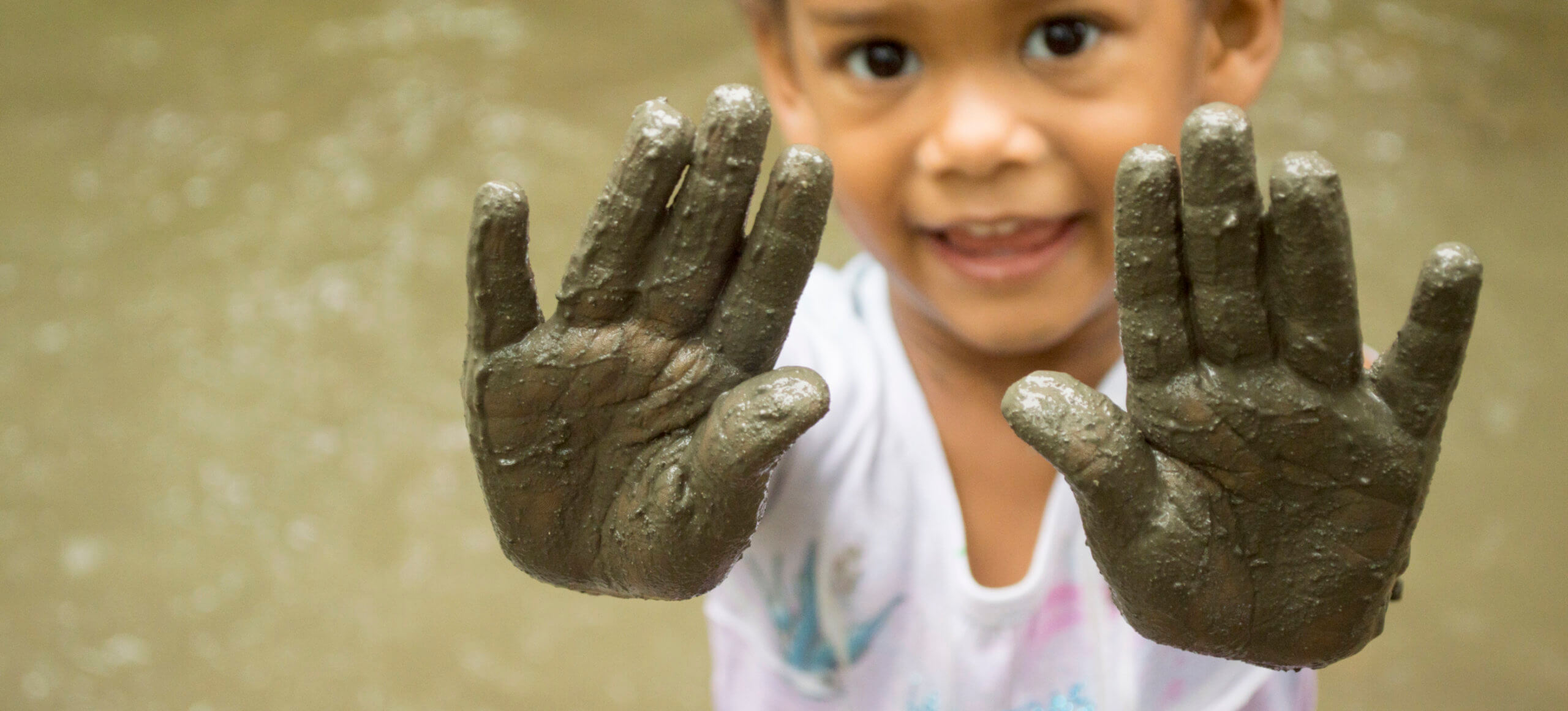 A child smiles in front of water while holding up her muddly palms.