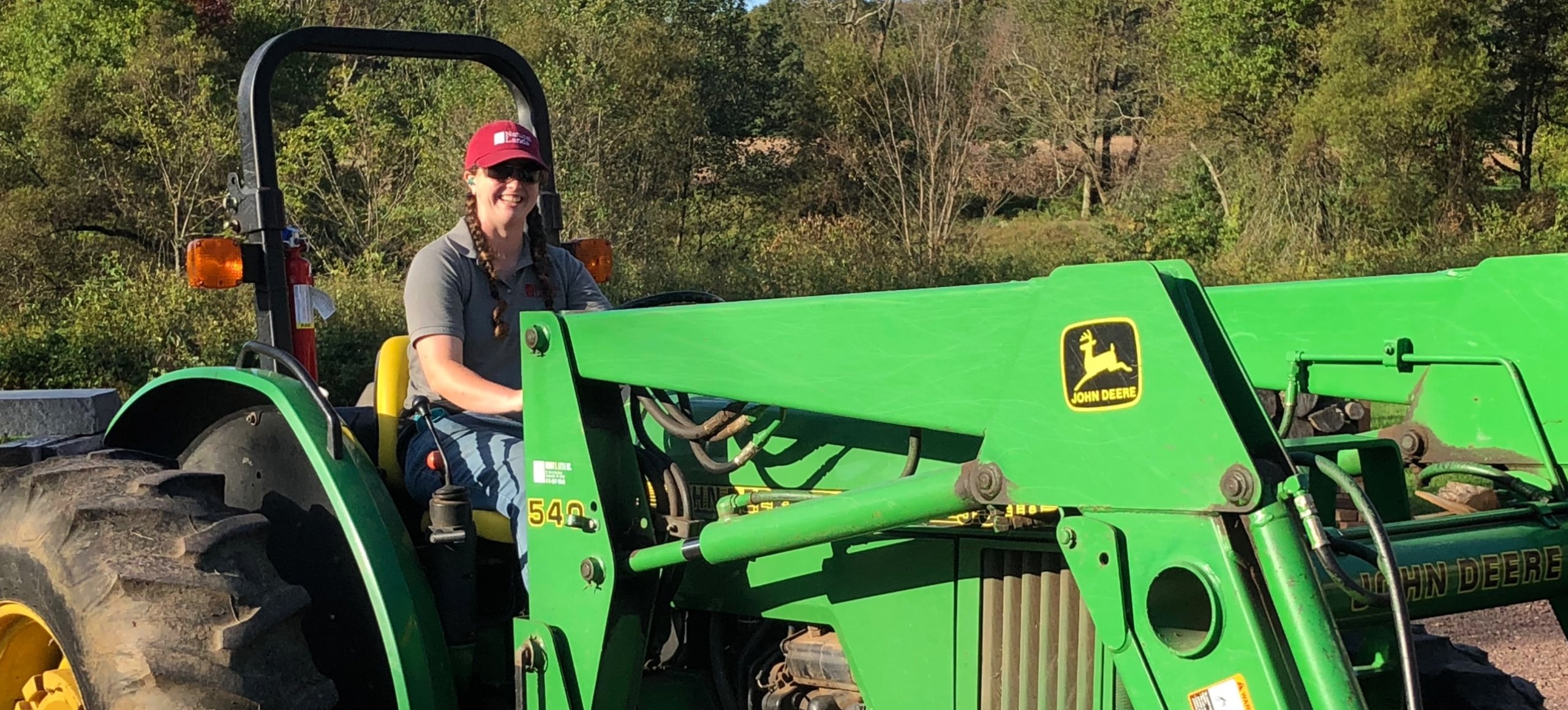 A person wearing a red ballcap sits in a large green tractor outside, smiling at the camera.
